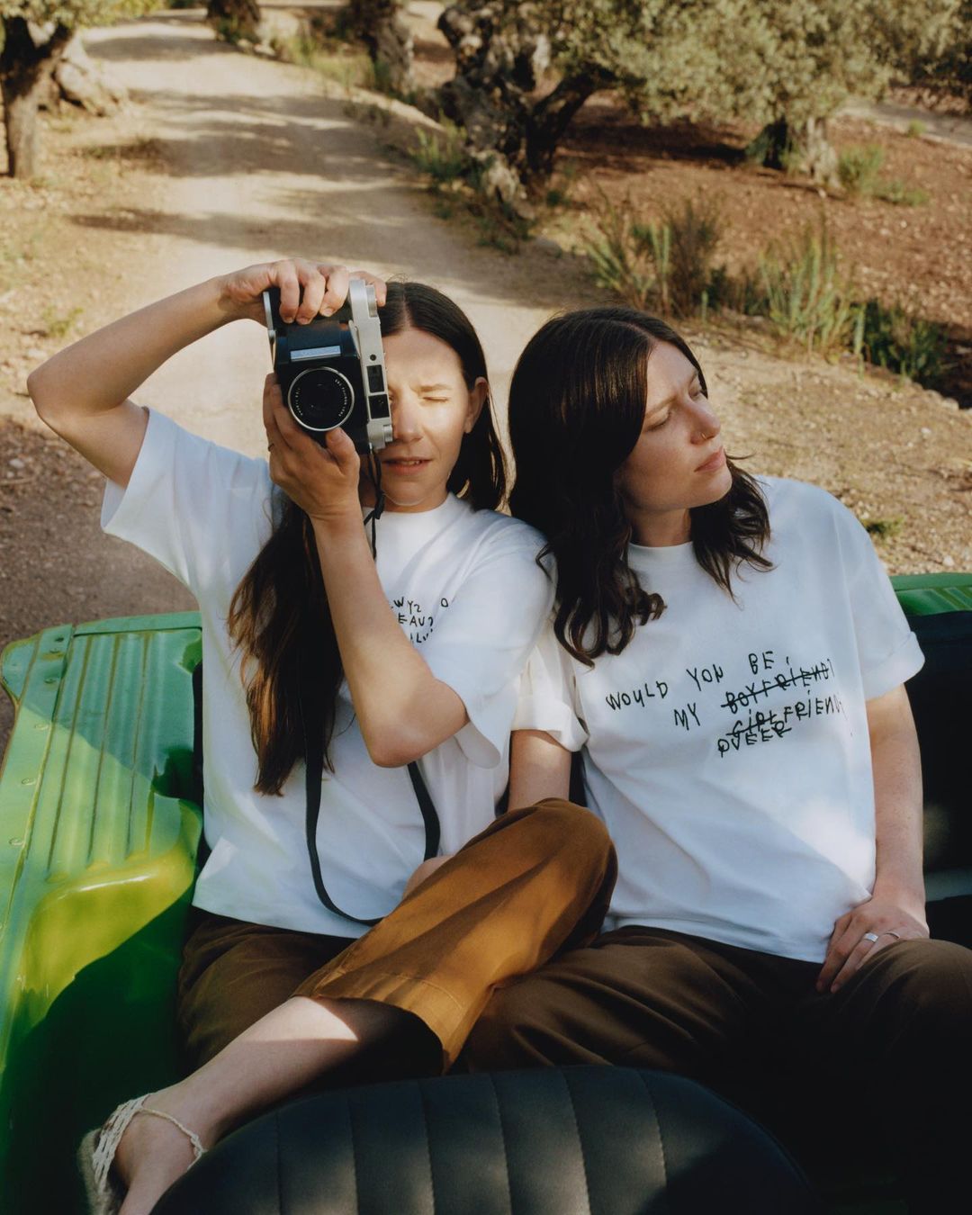 Two women in a green vehicle, one holding a camera. T-shirts feature handwritten text. Trees and a dirt path form the backdrop.