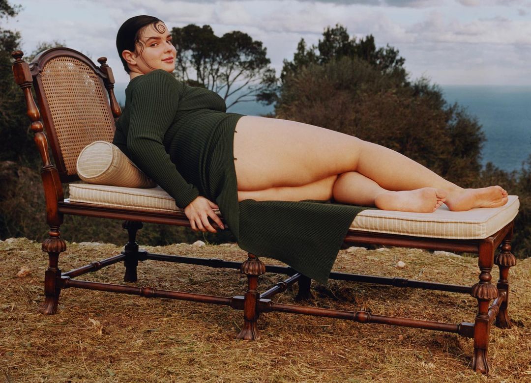 Adult woman poses on a wooden chair outdoors with trees, ocean and sky in the background. She is wearing a long-sleeved green dress.