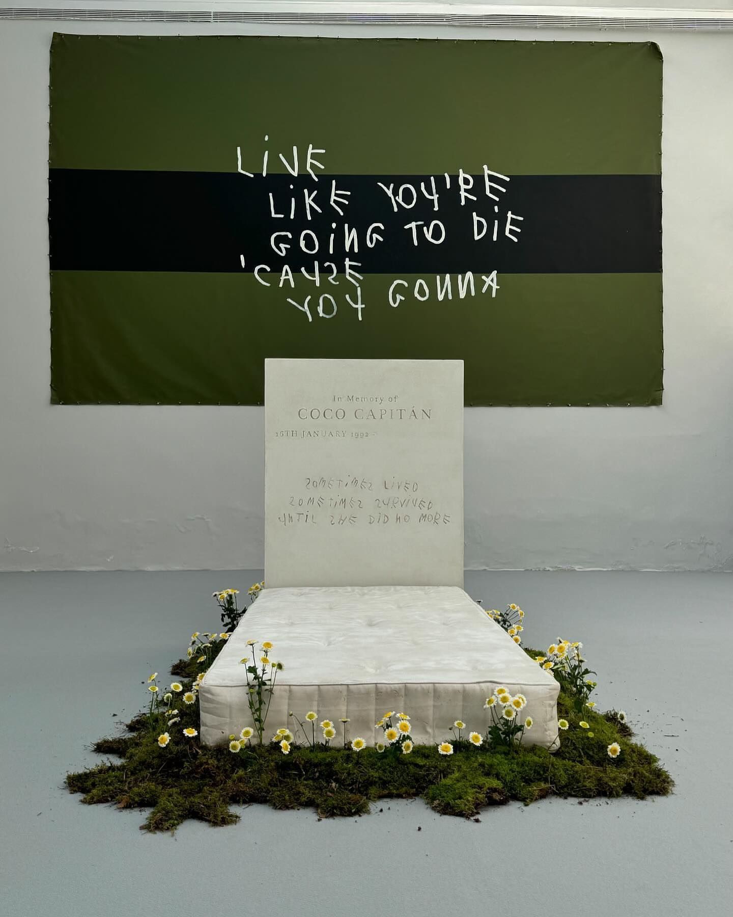 Conceptual art installation featuring a bed with floral arrangements, a tombstone-like headboard, and a motivational banner hanging above.