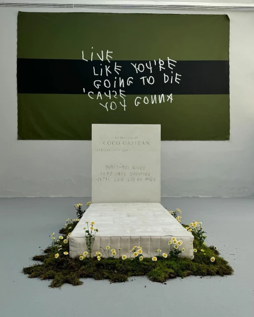 Conceptual art installation featuring a bed with floral arrangements, a tombstone-like headboard, and a motivational banner hanging above.