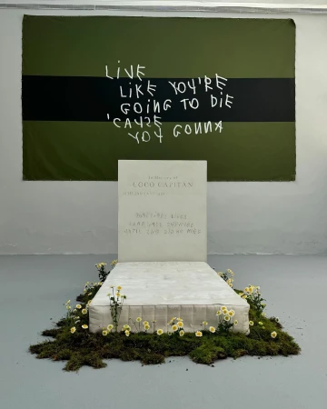 Conceptual art installation featuring a bed with floral arrangements, a tombstone-like headboard, and a motivational banner hanging above.