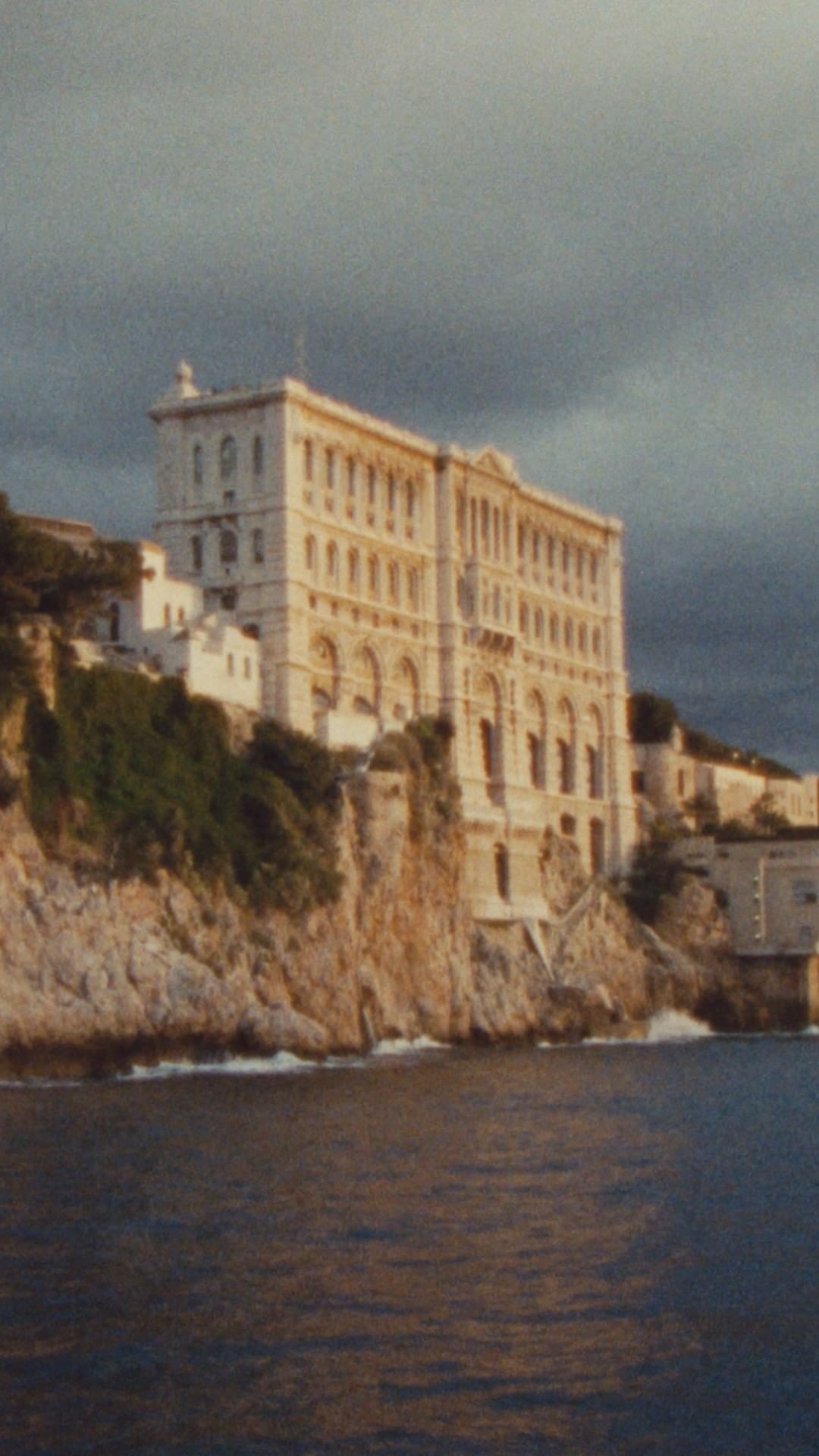 A grand neoclassical building perched atop a rocky cliff edge overlooking the sea under a dark stormy sky.