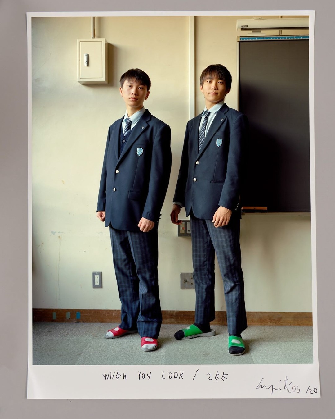 Two Asian teenage boys in school uniforms stand indoors against a neutral wall with a blackboard. Studio lighting highlights their attire.