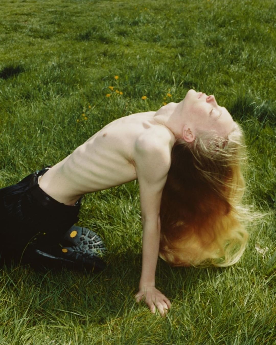 A young adult with long blonde hair arches their back while kneeling in a grassy field, with a bright and airy outdoor setting.