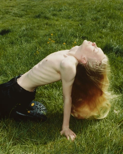 A young adult with long blonde hair arches their back while kneeling in a grassy field, with a bright and airy outdoor setting.