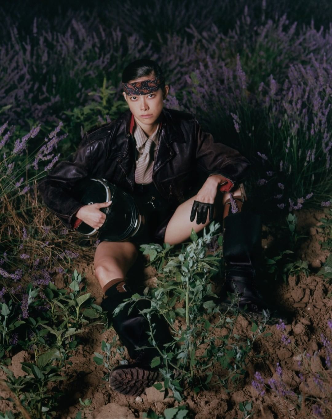 Asian male model in leather jacket seated among lavender flowers, holding helmet with black gloves and boots; night photography in field.