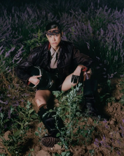 Asian male model in leather jacket seated among lavender flowers, holding helmet with black gloves and boots; night photography in field.