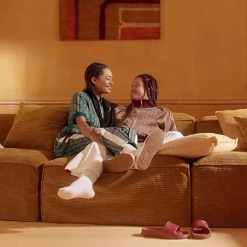 Two women sit together on a corduroy couch in a relaxed living room, interacting, dressed in casual clothing with natural light and muted colors.