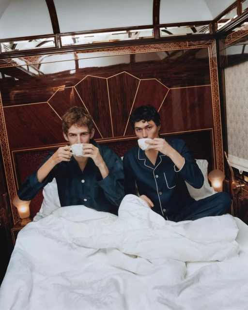 Two adult males in pajamas are sitting in bed, each drinking from a teacup, against an ornate wooden headboard and luxury bedding.