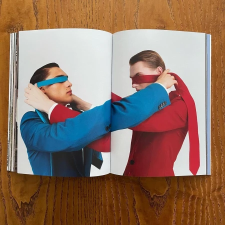 An open magazine spread on a wooden table displays portraits of two men blindfolded with colorful fabric ties.