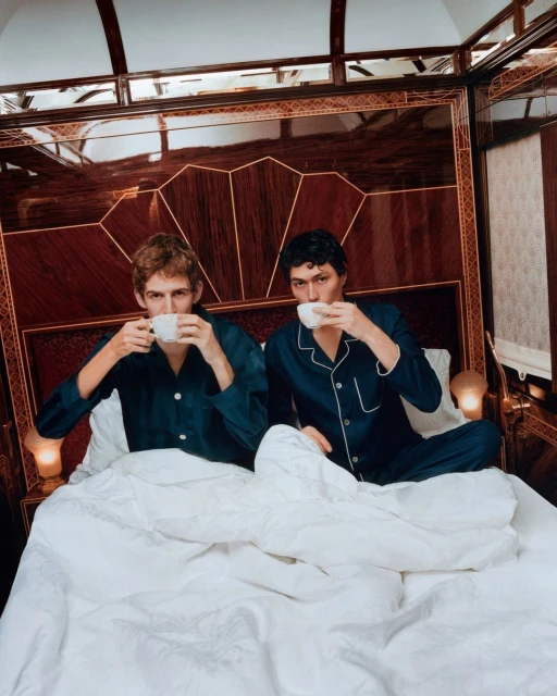 Two men in pajamas sit in a luxurious train bed drinking from teacups, set against dark wood paneling and white linens.