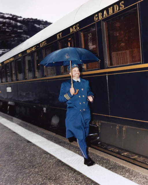 Adult man in uniform runs on train platform with umbrella next to train with gold lettering and mountain range in the background.