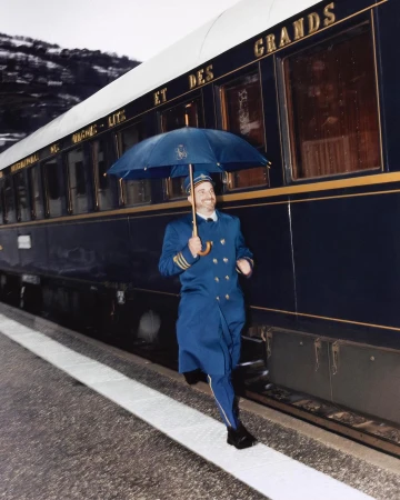 Adult man in uniform runs on train platform with umbrella next to train with gold lettering and mountain range in the background.