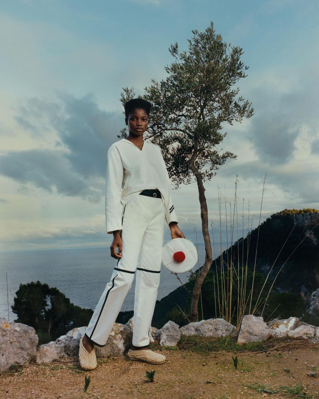 Fashion shoot of Black woman in cream-colored pantsuit holding a sun hat, set against a sky, sea, and tree landscape.