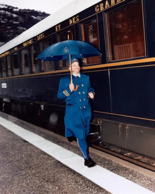 Man in blue uniform and umbrella running on train platform alongside a vintage train with snow-capped mountains in background.