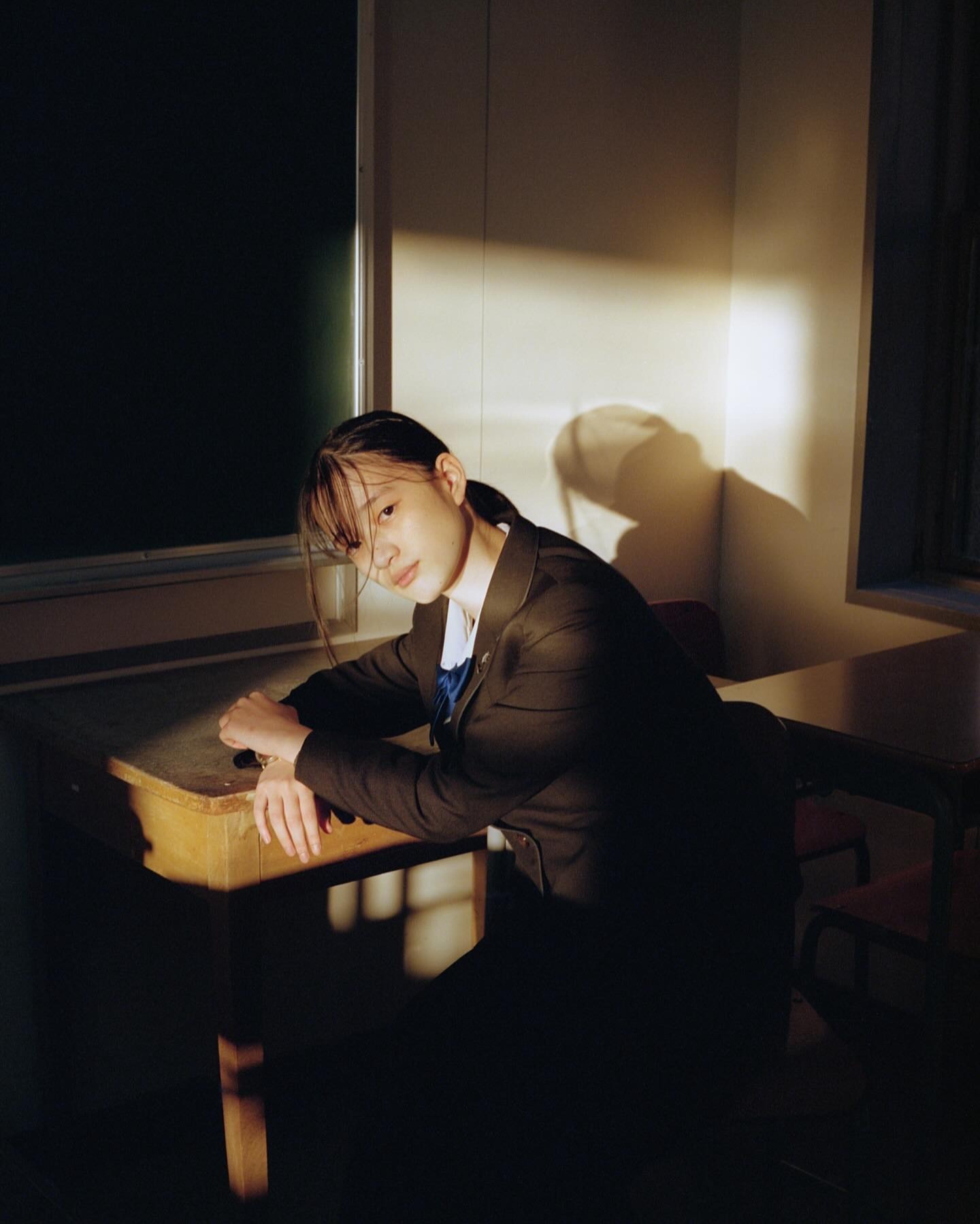 Young Asian woman poses indoors in school uniform, sitting at a desk with natural light casting shadows on wall in the background.