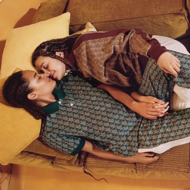 A couple reclines on a yellow couch, their faces close, wearing matching patterned polo shirt and sweatshirt, enjoying an intimate moment indoors.