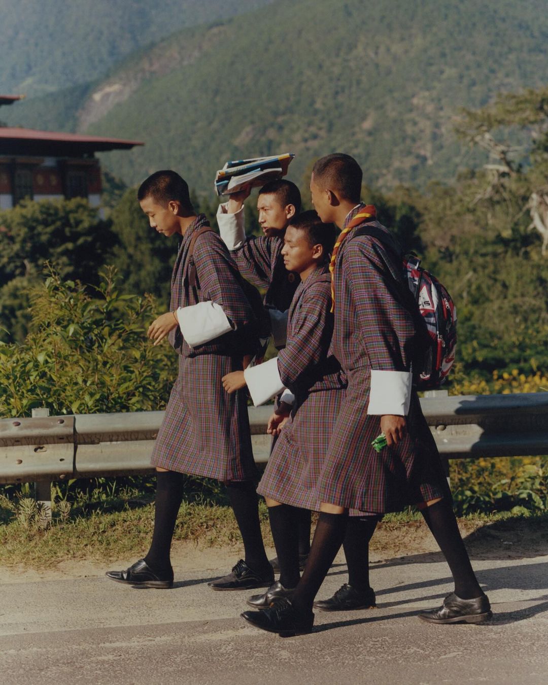Four young Bhutanese students in traditional uniforms walk on a road with books and a backpack, with mountains and buildings behind them.