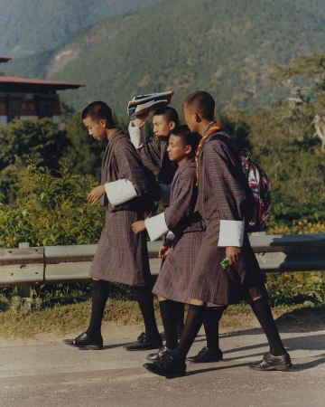 Four young Bhutanese students in traditional uniforms walk on a road with books and a backpack, with mountains and buildings behind them.