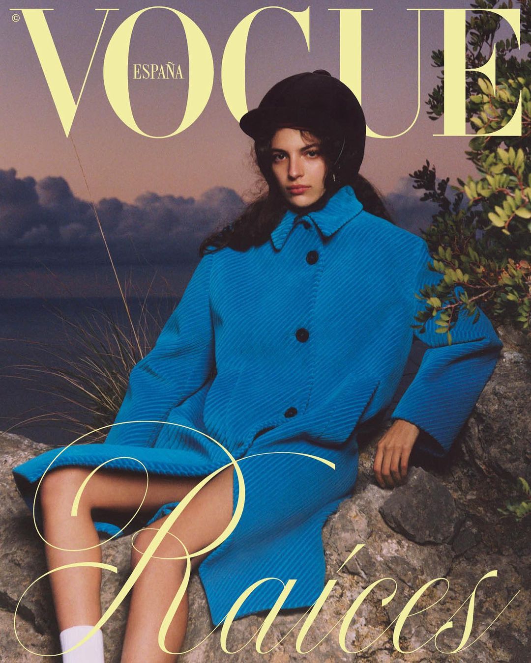 Vogue España cover with a woman in a blue coat and black hat posing outdoors against a cloudy sunset sky and ocean backdrop.