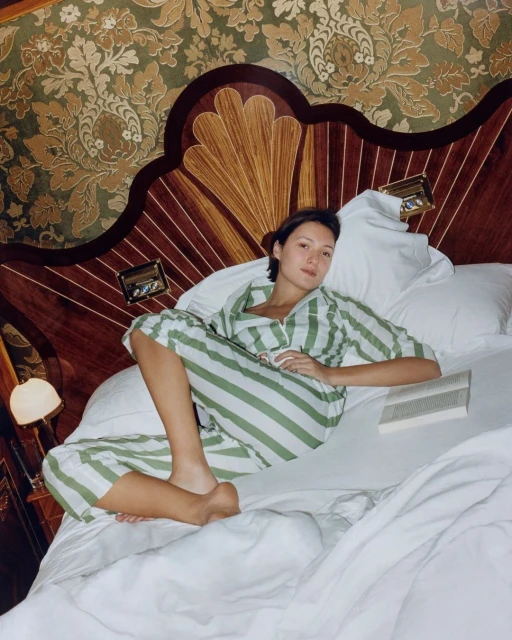 Adult woman in striped pajamas lounging on a bed with white linens in an indoor bedroom setting with patterned wallpaper.