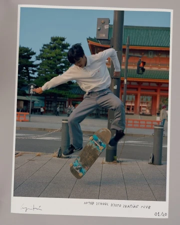 Asian male skateboarder mid-air in front of traditional Japanese architecture, wearing a white shirt and grey pants, urban environment.