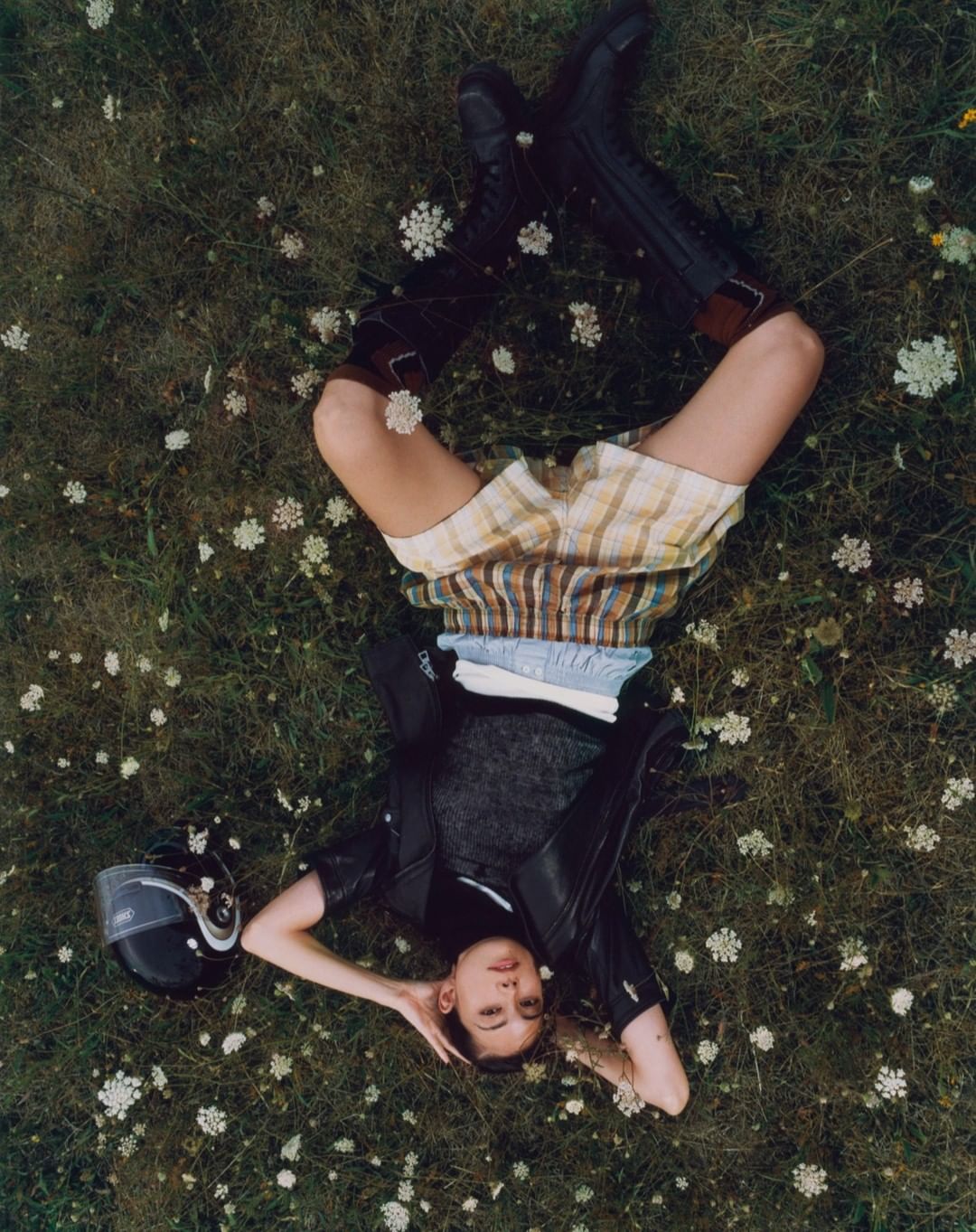 Woman in shorts and boots lies on green grass with white flowers, wearing a leather jacket next to a black motorcycle helmet, overhead shot.