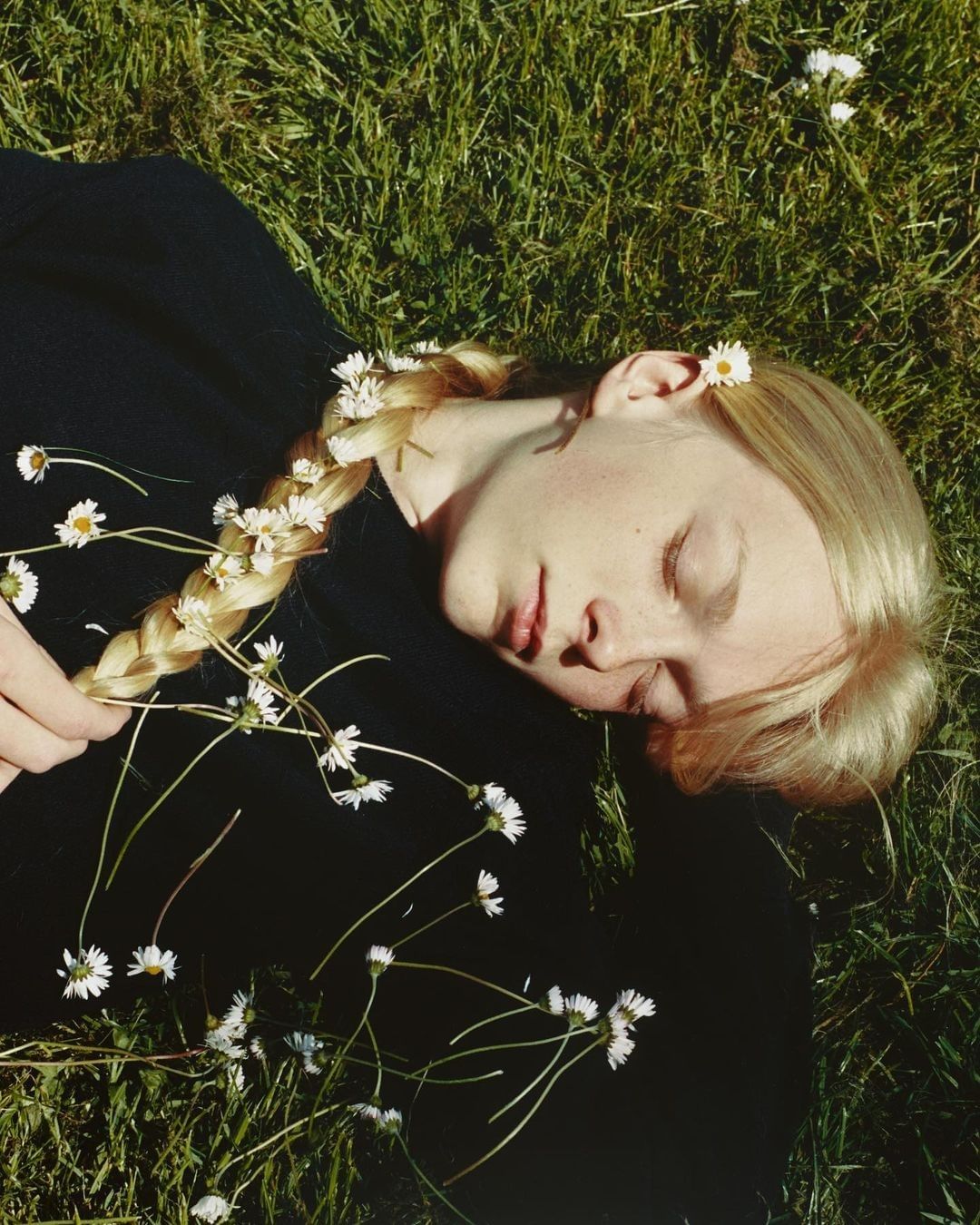 Tranquil scene of a fair-skinned woman with a blonde braid adorned with daisies lying in vibrant green grass, eyes closed, natural light.