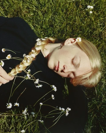 Tranquil scene of a fair-skinned woman with a blonde braid adorned with daisies lying in vibrant green grass, eyes closed, natural light.