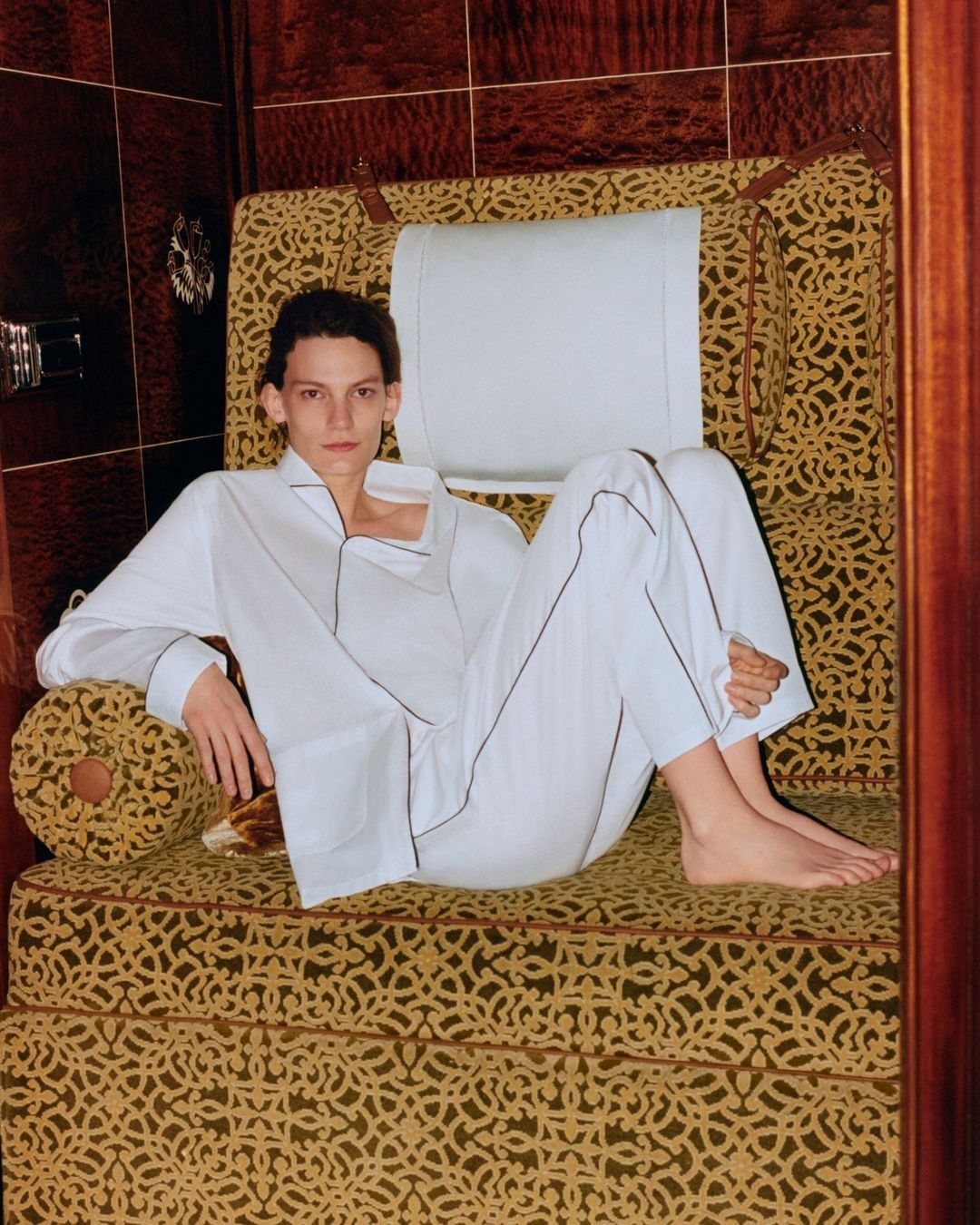 An adult poses on an upholstered chair in white pyjamas with black piping, inside a room with dark brown wood-paneled walls.