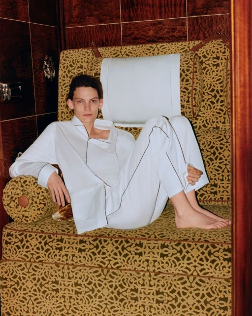 An adult poses on an upholstered chair in white pyjamas with black piping, inside a room with dark brown wood-paneled walls.