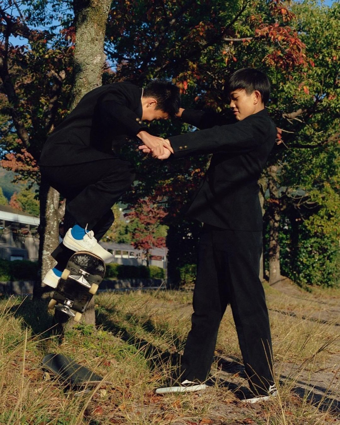 Two male teenagers in school uniforms balancing on a skateboard in an outdoor setting with autumn foliage and buildings visible in the background.