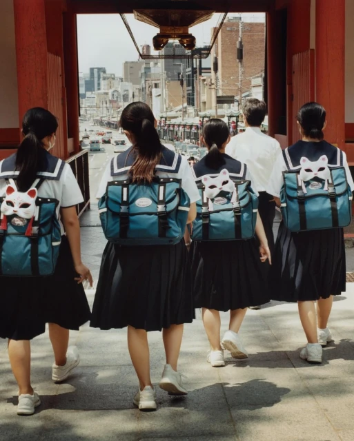A group of students in school uniforms walk away from the camera, entering a city street through a red wooden temple gate.