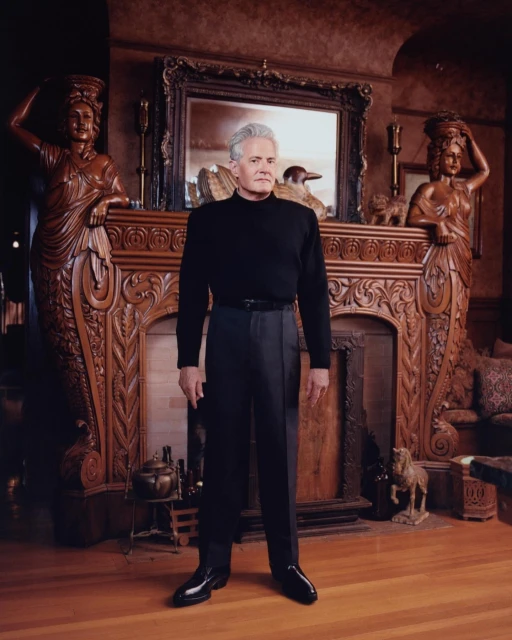 A man stands in front of an ornate fireplace in an opulent room featuring carved statues, dark wood, and a large decorative mirror.