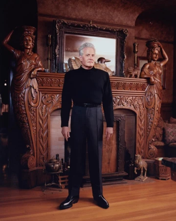 A man stands in front of an ornate fireplace in an opulent room featuring carved statues, dark wood, and a large decorative mirror.
