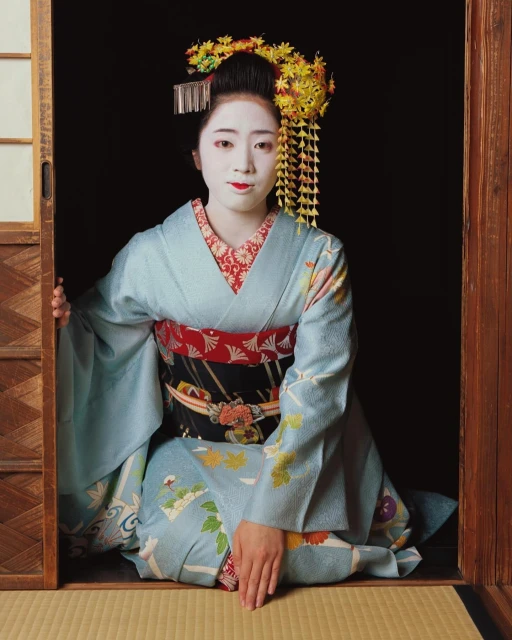 Woman in traditional Japanese geisha attire with white face makeup, elaborate yellow hair ornaments, and a light blue kimono.