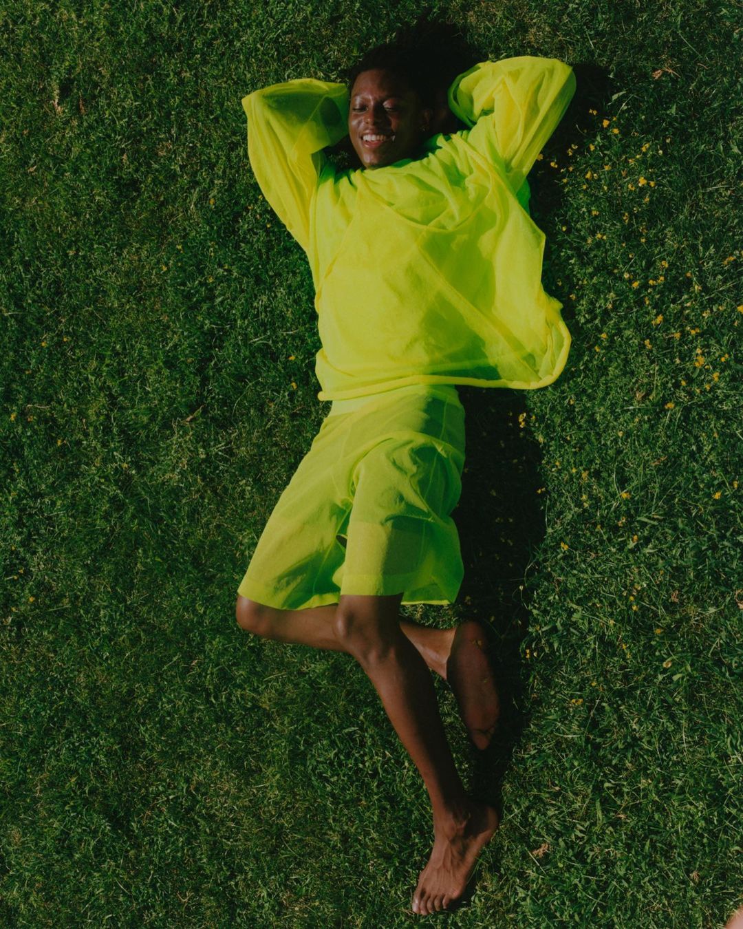 Person lies on green grass wearing matching neon-yellow top and shorts, relaxing with hands behind head, smile, and bare feet.
