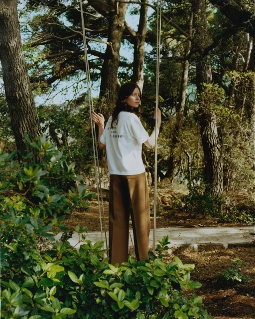 Woman on a swing in an outdoor forest setting, wearing a white t-shirt and brown pants surrounded by lush green foliage.