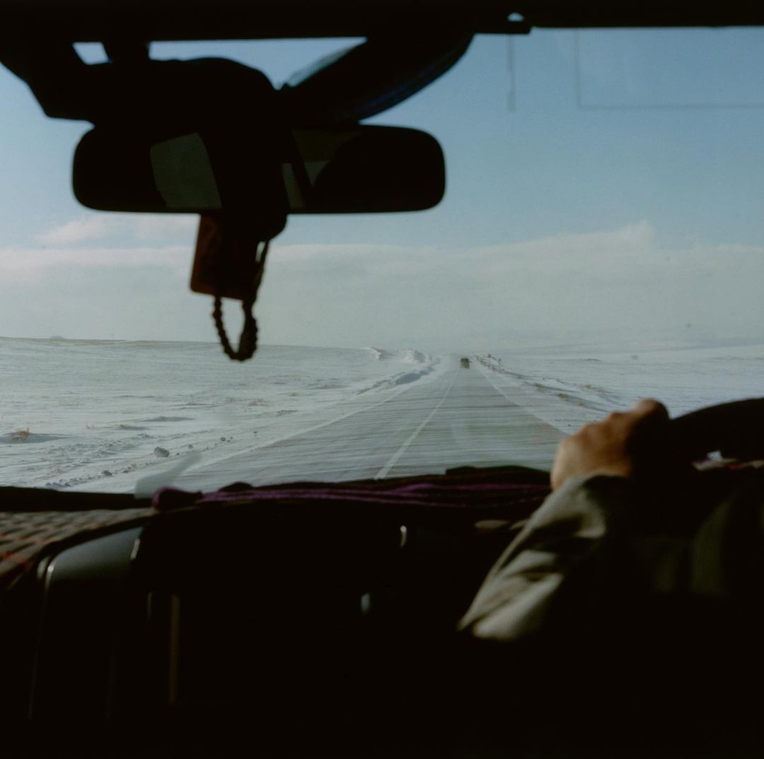 Road trip view from inside a car: icy highway stretches through a flat, snowy landscape under a blue sky with scattered clouds.