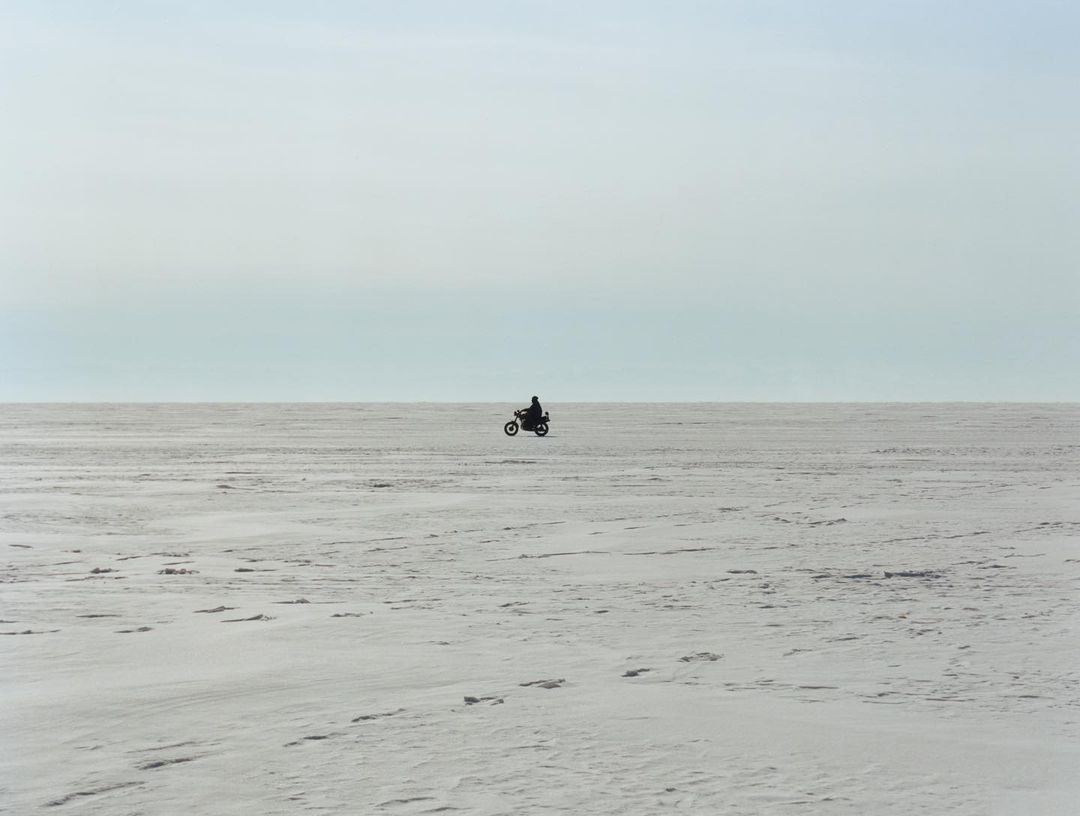 A motorcycle rider silhouetted in the center of a snowy, flat expanse under a bright sky with soft, diffused lighting.