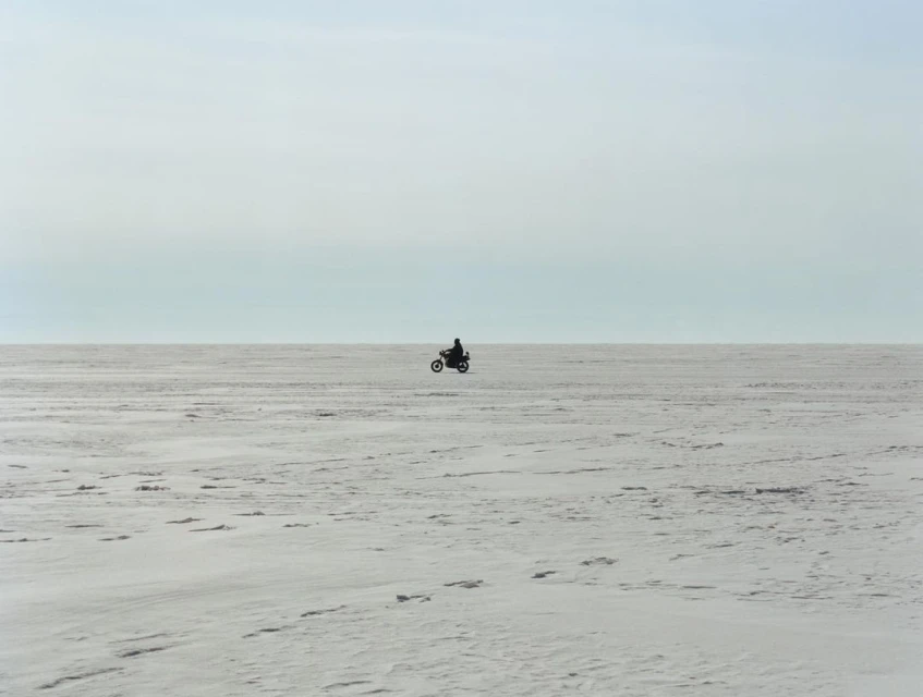 A motorcycle rider silhouetted in the center of a snowy, flat expanse under a bright sky with soft, diffused lighting.