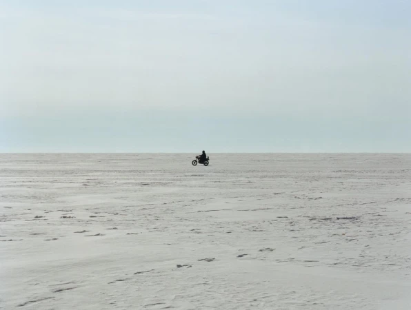 A motorcycle rider silhouetted in the center of a snowy, flat expanse under a bright sky with soft, diffused lighting.