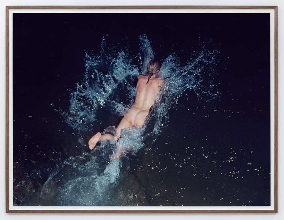 Nude adult female plunges backwards into dark water with splash, photographed from above, framed with white matte and dark brown wood.