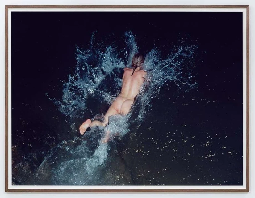 Nude adult female plunges backwards into dark water with splash, photographed from above, framed with white matte and dark brown wood.