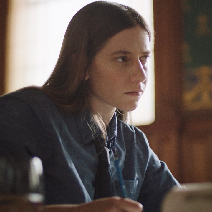 Adult female with long brown hair is sitting at a table writing in an indoor environment, wearing a button-down shirt and dark tie.