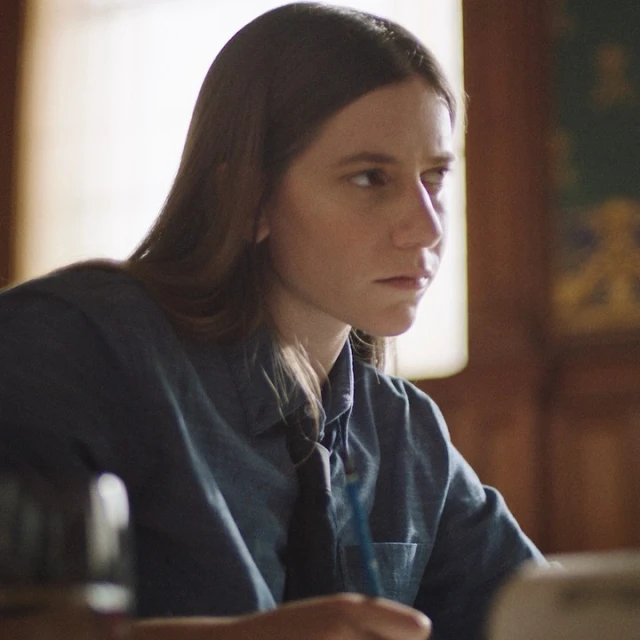 Adult female with long brown hair is sitting at a table writing in an indoor environment, wearing a button-down shirt and dark tie.