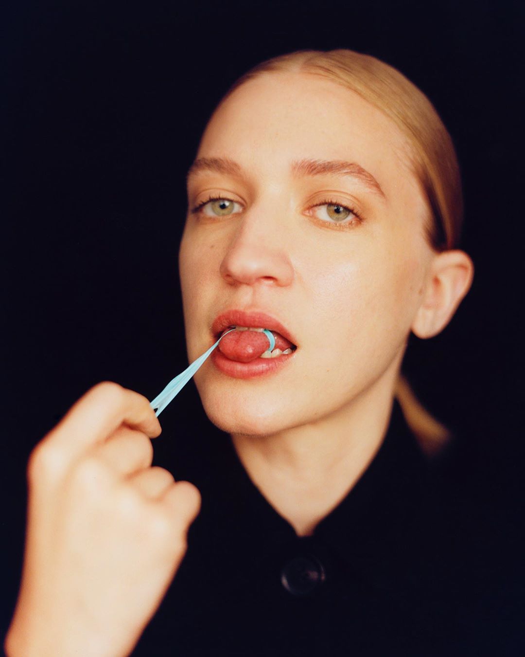 Close-up of an adult woman with fair skin holding a blue dental pick, cleaning her tongue in a studio setting with a black backdrop.