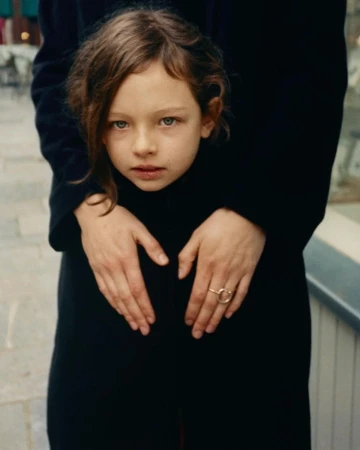 A child framed by an adult wearing a black coat with hands resting on the child's chest, photographed by Coco Capitan.