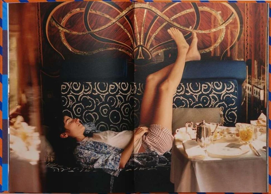 A woman reclines on a patterned sofa next to a breakfast table, photographed for an editorial spread in natural light with a decorative backdrop.