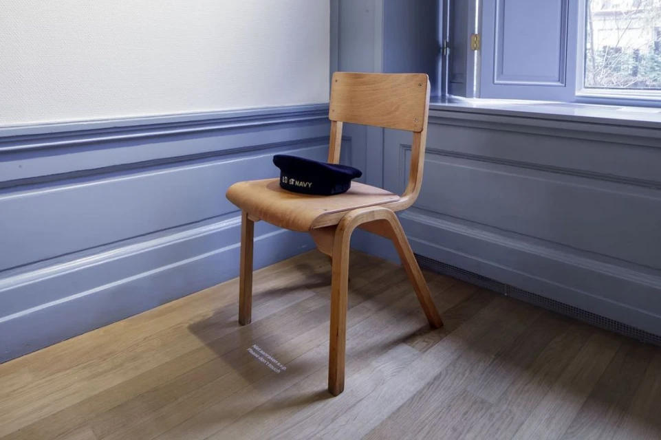 A wooden chair with a navy hat on its seat sits indoors on a wooden floor with a view of trees through the window in the background.